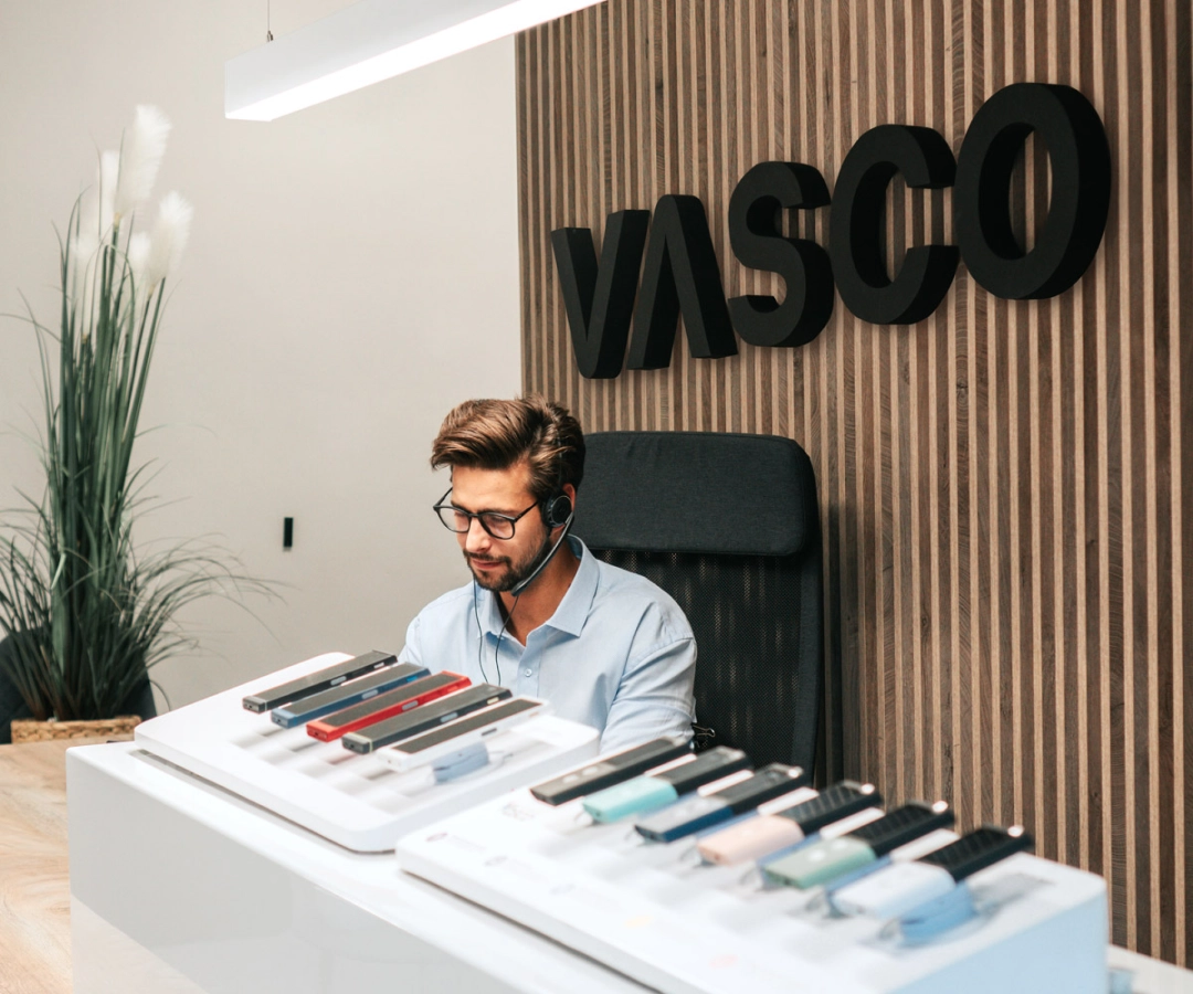 Čovjek iz korisničke službe sjedi za stolom koji prikazuje više Vasco Translator uređaja u raznim bojama, s velikim natpisom 'VASCO' na drvenom zidu iza njega.