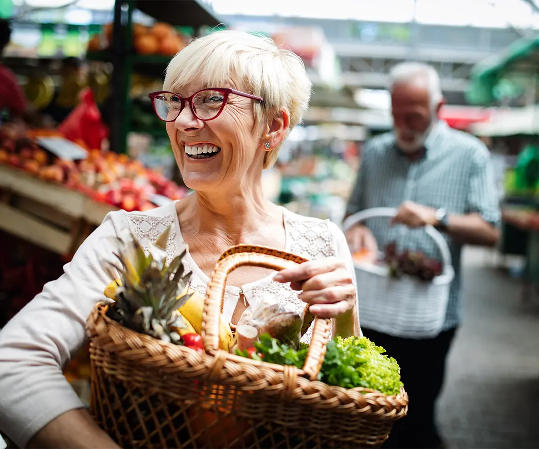 A smiling older woman with short blonde hair and red glasses carrying a wicker basket filled with fresh vegetables and fruit at an outdoor market. In the blurred background, another person is shopping among stalls of produce.